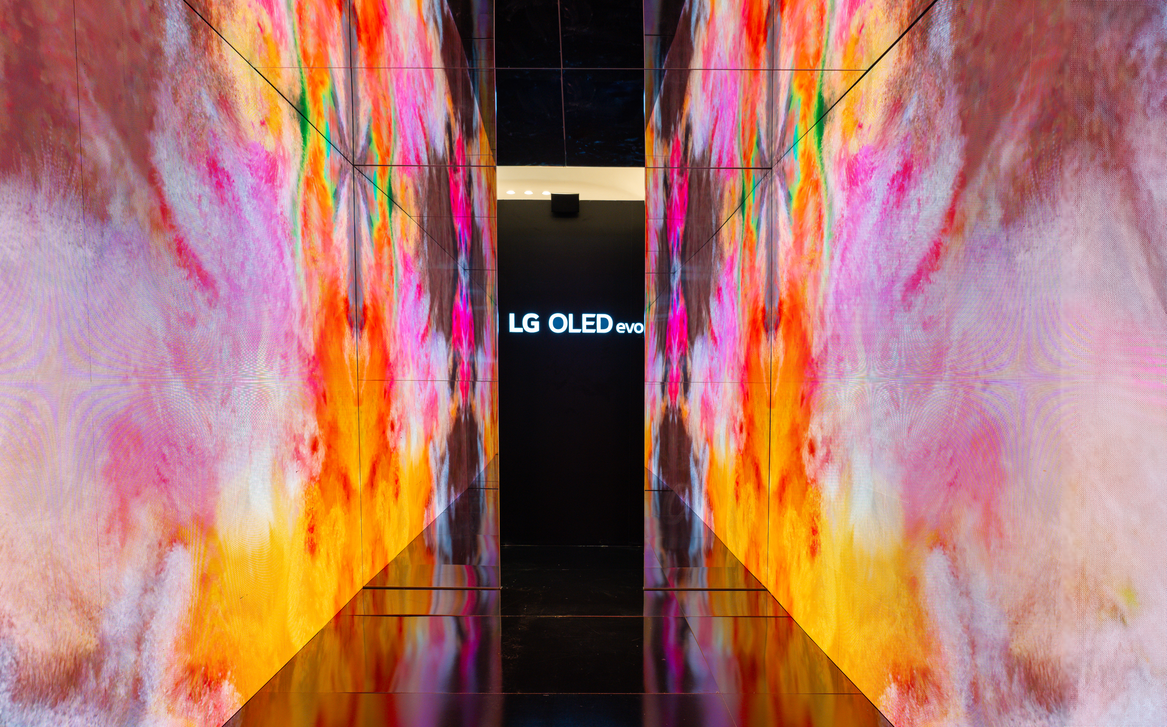 The immersive wall displaying an alternate generative installation. The screens show textured patterns in warm tones of pink, orange, and yellow, reflecting across the dark floor toward the central signage.